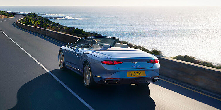 Bentley Tallinn New Bentley Continental GTC Azure convertible rear view in Light Windsor blue paint with 22 inch ten spoke wheels driving dynamically on an ocean road in the sunshine