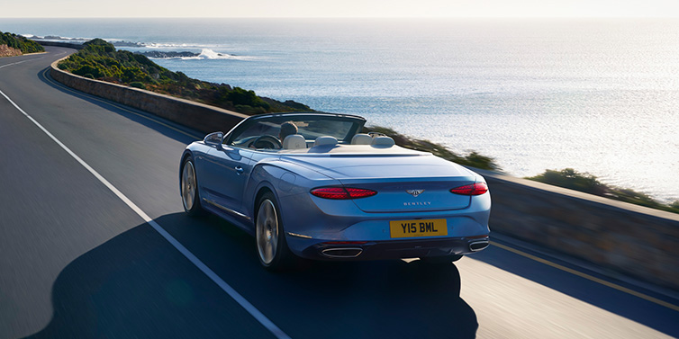 Bentley Tallinn New Bentley Continental GTC Azure convertible rear view in Light Windsor blue paint with 22 inch ten spoke wheels driving dynamically on an ocean road in the sunshine